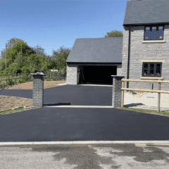 A newly paved black driveway, featuring expert driveway installation, leads to a stone house with a garage. The path is flanked by wooden and stone fences. Greenery and trees adorn the background under a clear blue sky.