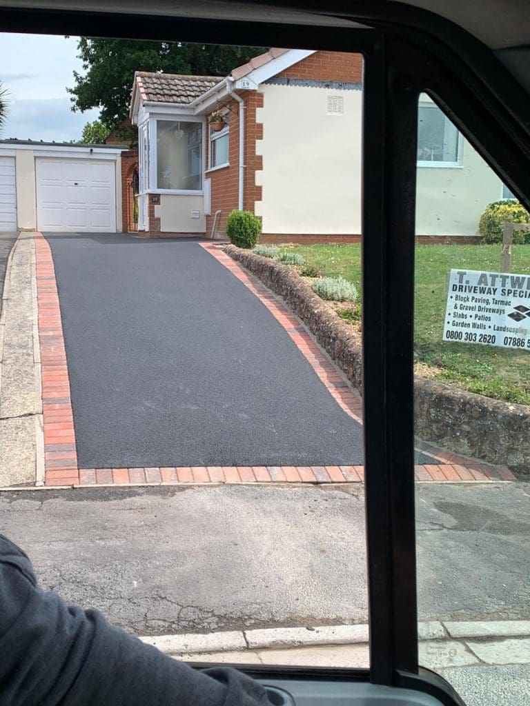 Through the car window, a freshly paved driveway leads to a garage attached to a brick house. A sign on the lawn reads "Driveway Specialist" with contact information. The red brick edging highlights expert Driveway Installers work, perhaps specializing in Resin Driveways for a sleek finish.