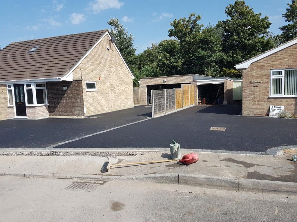 A suburban scene with two brick bungalows and freshly laid resin bound driveways. The house on the left flaunts a gabled roof while the one on the right features a flat-roofed garage. A broom, dustpan, and bucket rest on the sidewalk after a driveway installation in the foreground.