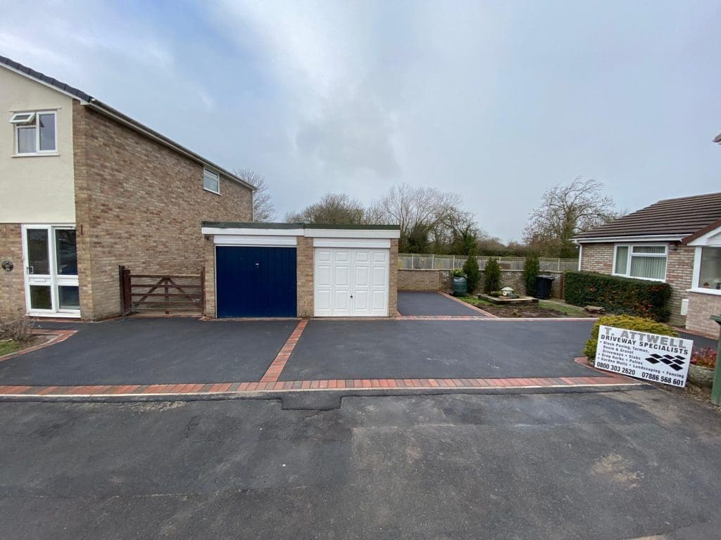 A freshly laid tarmac driveway connects to a brick house and garage, featuring one blue door and one white door. A sign reading "B. Atwell Driveway Specialists" sits on the right, set against resin-bound pathways, with trees and a cloudy sky adding charm in the background.