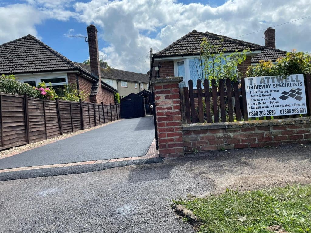A newly paved resin bound driveway leads to a single-story brick house. Two similar houses are on either side, separated by brown wooden fencing. A sign on the right advertises driveway services with a phone number. The sky is partly cloudy.