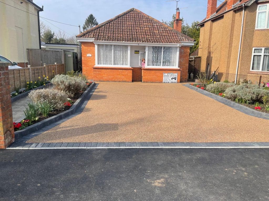 A small brick house with a tiled roof features a wide, tan-colored driveway expertly crafted by skilled driveway installers, bordered by flower beds. The flower beds are lined with gray edging stones and filled with plants and colorful flowers. The house has a central door and large front windows.