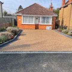 A small brick house with a tiled roof features a wide, tan-colored driveway expertly crafted by skilled driveway installers, bordered by flower beds. The flower beds are lined with gray edging stones and filled with plants and colorful flowers. The house has a central door and large front windows.