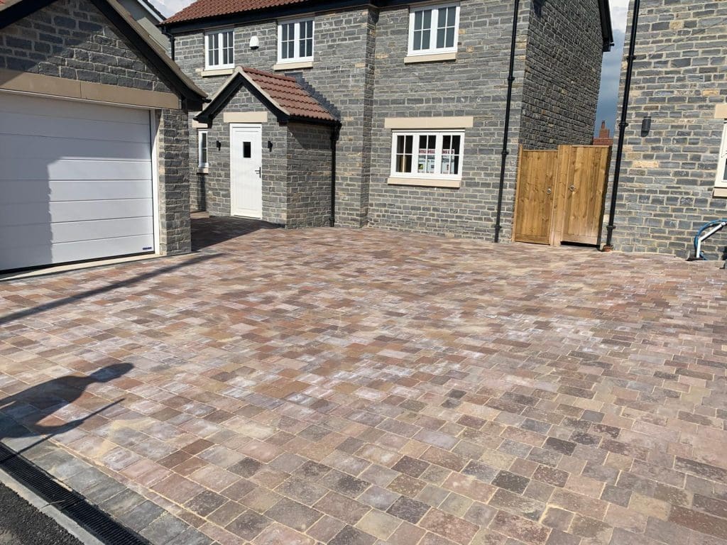 A block paving driveway leads to a two-story stone house with a dark brown roof. There is a white garage door to the left and a wooden fence to the right. The house features white-framed windows and a white front door, all under a partly cloudy sky.