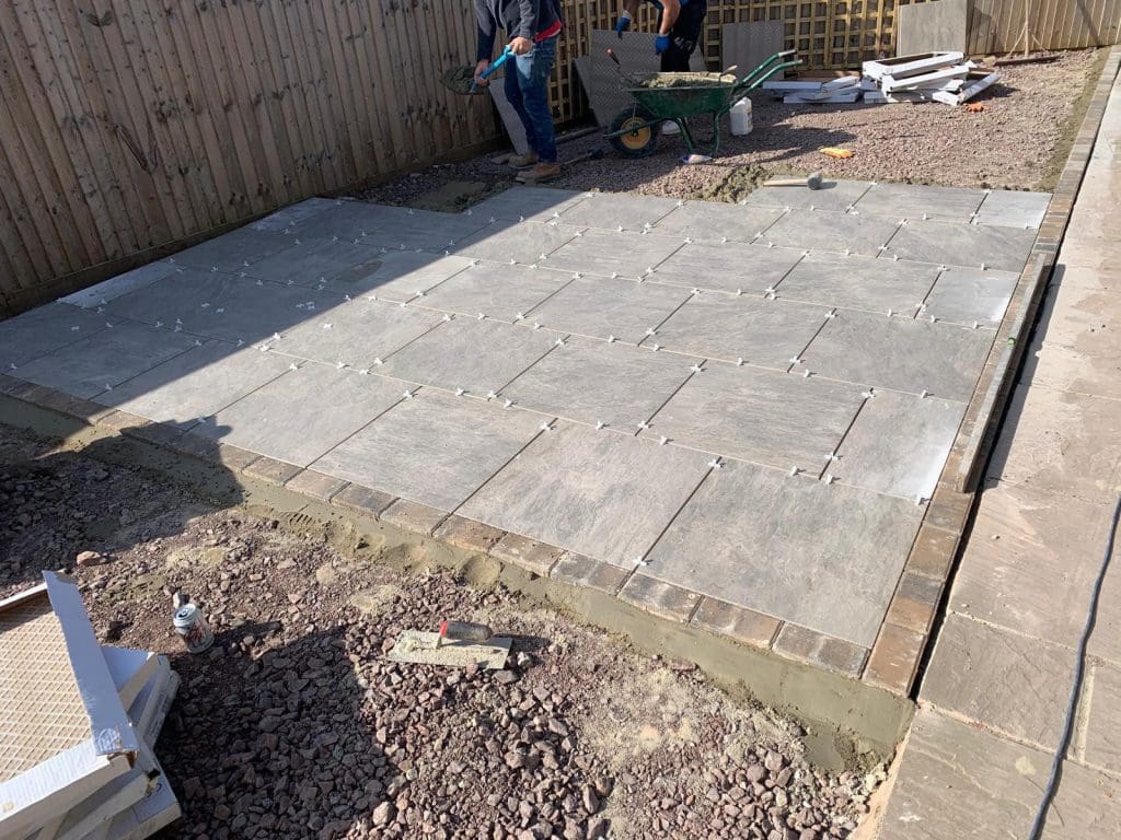Two people are laying large gray stone tiles on a patio, similar to the expertise of skilled driveway installers. Some tiles are already placed, with spacers visible. A wheelbarrow with tools is nearby, and a wooden fence runs along one side of the yard.