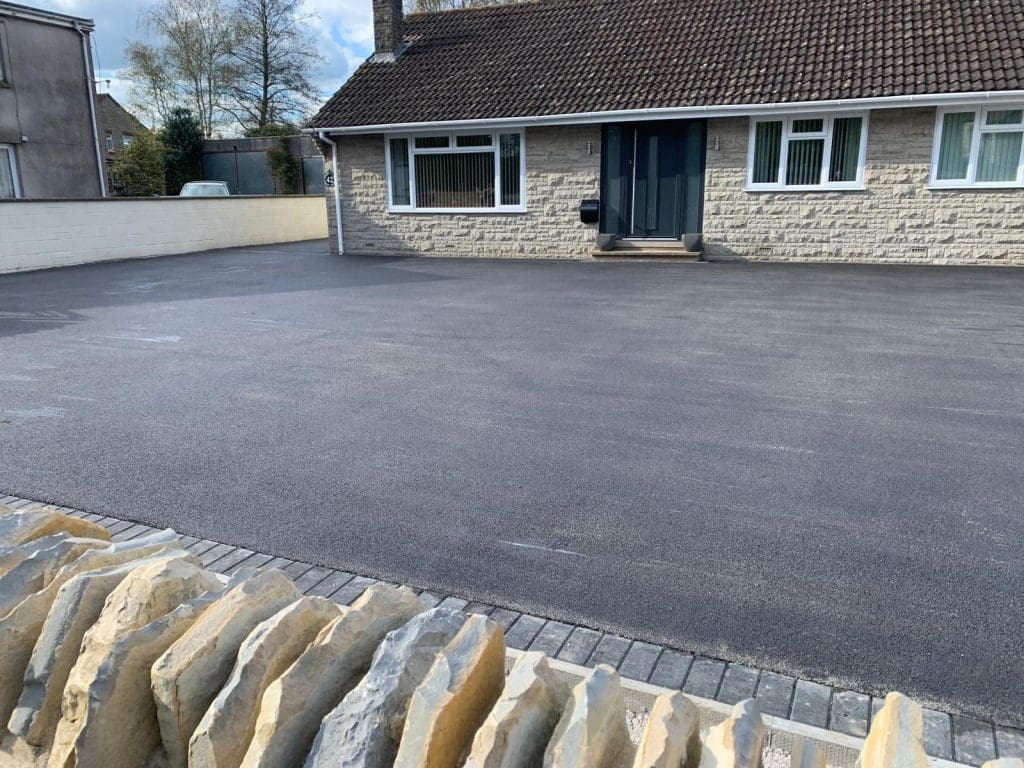 A newly installed, expansive asphalt driveway graces the front of a charming single-story house with a tiled roof. The stone facade, accented by white-framed windows and a dark blue door, exudes elegance. In the foreground, a decorative stone wall adds character, with trees providing a natural backdrop.