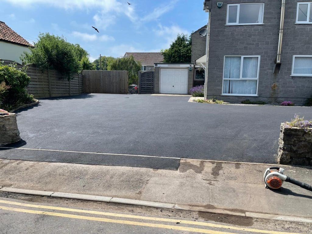 Freshly paved driveway in front of a gray stone house with a white garage door, featuring beautiful block paving. A leaf blower rests on the sidewalk beside the driveway. Blue sky with a few birds above. Garden beds with flowers and shrubs line the sides.