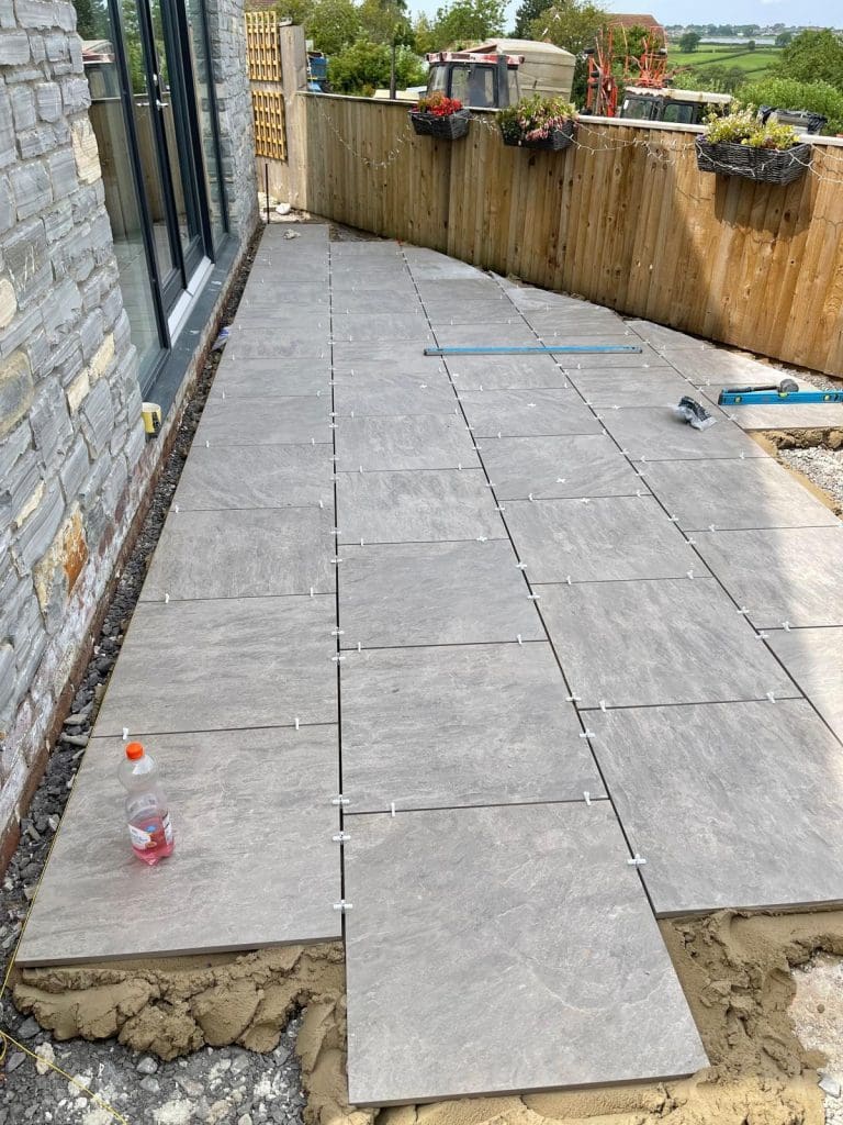 A patio under construction features large gray tiles being laid on a bed of sand. A stone wall stands to the left, with a wooden fence and flower pots on the right. Tools and a drink bottle are scattered among the tiles, similar to those used by block paving experts. The garden and sky stretch into the distance.