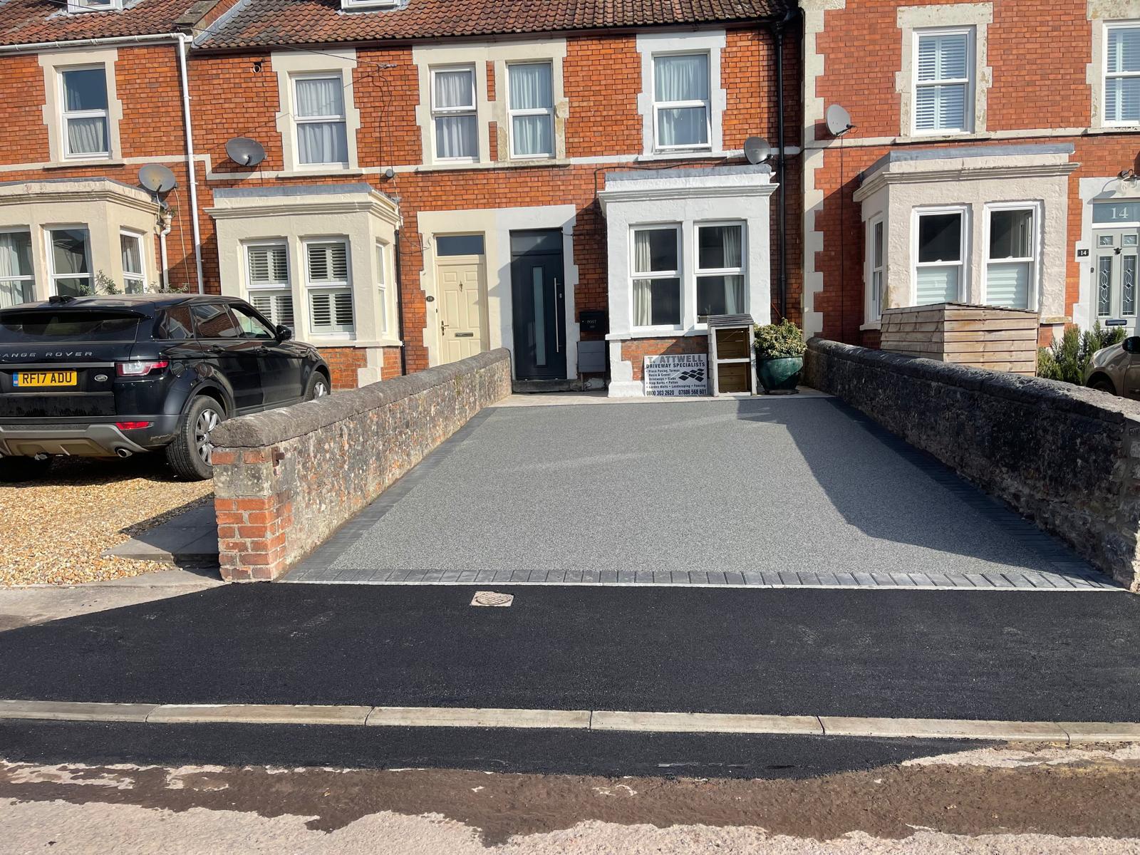A recently completed resin bond driveway installation leads to a brick townhouse with white-framed windows. A black car is parked on a neighboring gravel area. Brick walls and a neatly paved path frame the driveway, showcasing the expert work of skilled driveway installers.