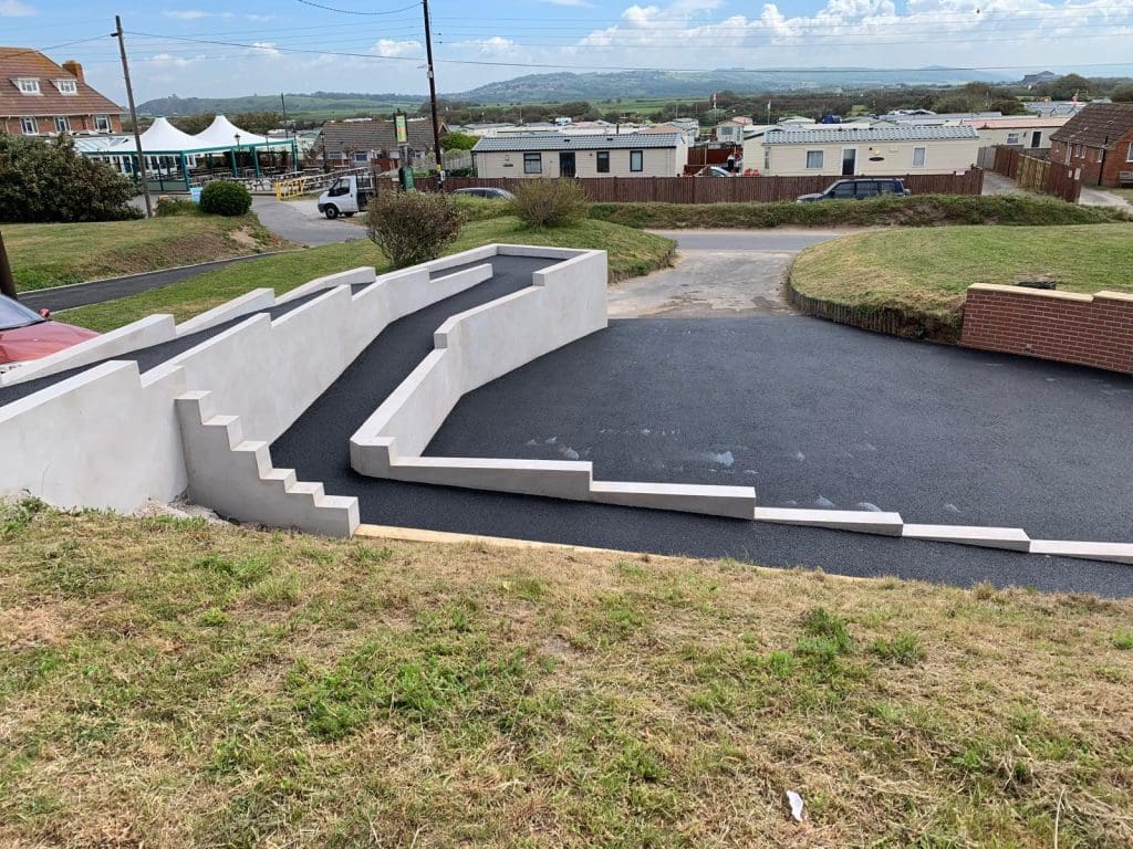 A newly constructed parking area with fresh asphalt and white painted walls showcases expert driveway installation. There are stairs on the left leading down to the area, framed by houses, a road, and distant hills under a cloudy sky.