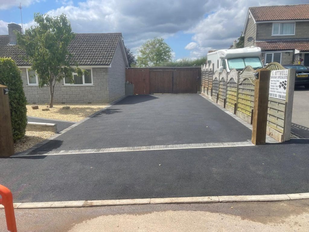 A newly paved driveway crafted by expert driveway installers graces the front of a single-story house. On the left, a tree and gravel landscaping complement the scene, while a parked van peeks from behind a wooden fence on the right. The sky is partly cloudy, adding to the serene setting.