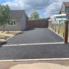 A newly paved driveway crafted by expert driveway installers graces the front of a single-story house. On the left, a tree and gravel landscaping complement the scene, while a parked van peeks from behind a wooden fence on the right. The sky is partly cloudy, adding to the serene setting.