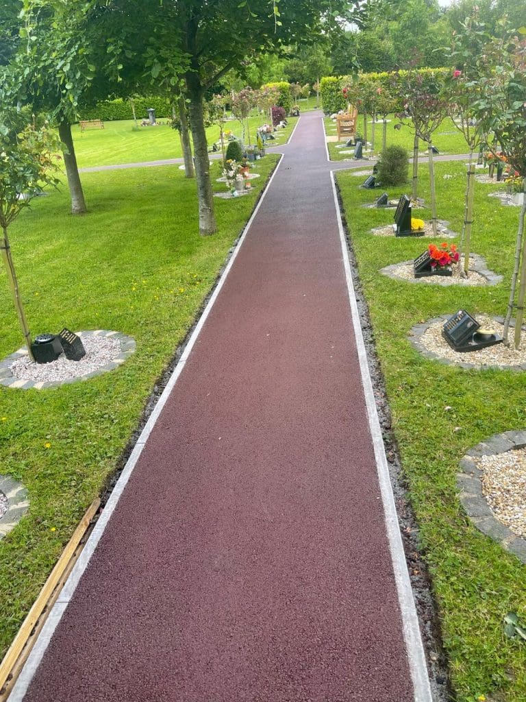 A narrow paved pathway bordered by grass leads through a serene garden, echoing the elegance of resin driveways. Trees and small memorial plaques with flowers align both sides, while benches are placed intermittently in the lush green setting.