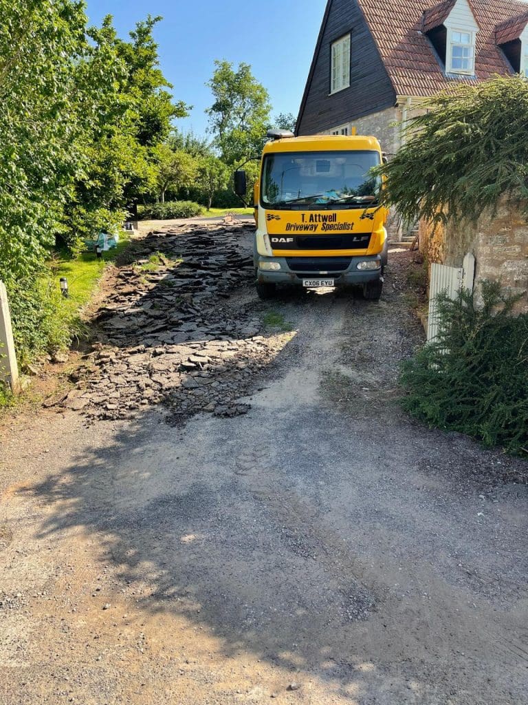A yellow truck parked on a gravel driveway in front of a house with a sloped tiled roof hints at upcoming changes. Broken pavement pieces are scattered around, while trees and shrubs border the area under a clear blue sky, awaiting skilled driveway installers to craft elegant resin driveways.