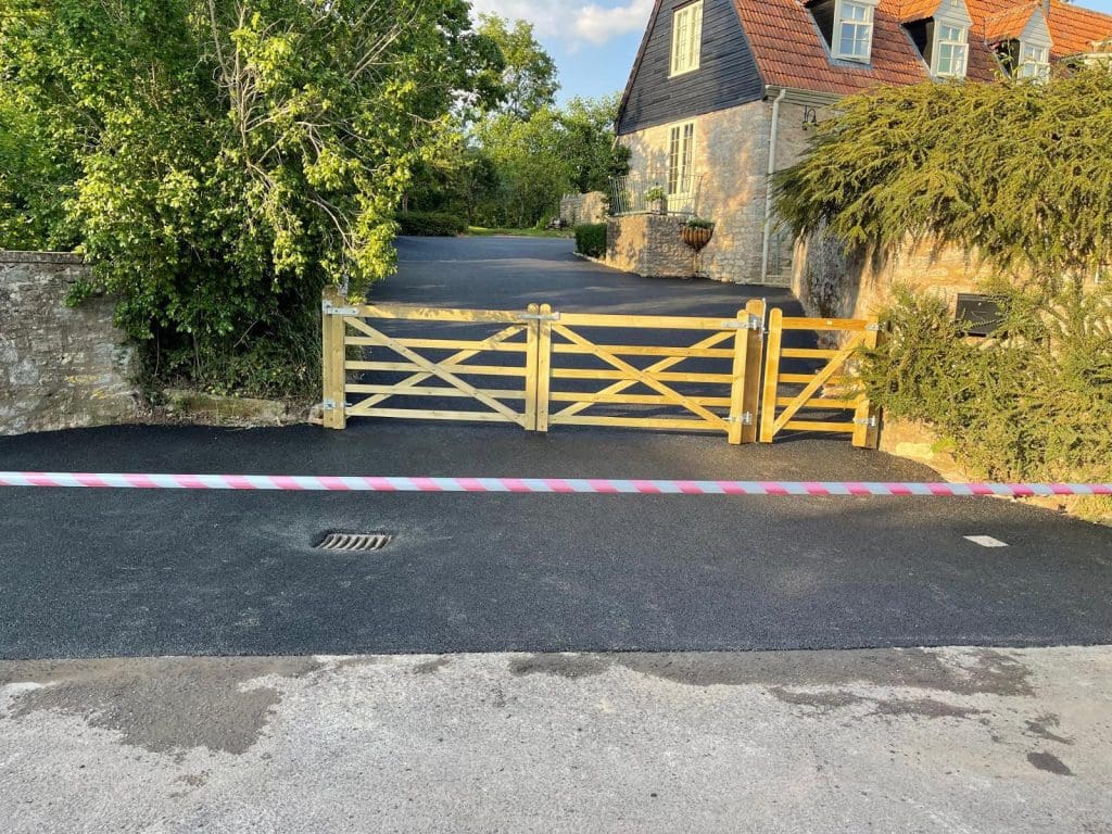 A wooden gate blocks a driveway leading to a stone house with a red-tiled roof. The freshly paved resin driveway is bordered by trees and a stone wall. A pink and white striped barrier is in the foreground, adding a touch of charm to the picturesque scene.