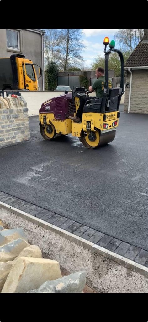 A person operates a yellow and purple road roller on freshly laid asphalt in a residential driveway, highlighting the expertise of skilled driveway installers. A parked truck and stone wall are visible in the background, along with some trees and a house.