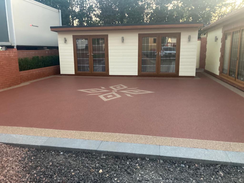 Spacious yard with a smooth, reddish-brown resin driveway featuring a geometric design. Two modern, white buildings with wooden accents have large glass doors. Surrounded by brick walls, bushes, and a paved edge.