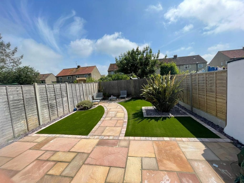 A backyard with a stone patio and curved pathway leads to artificial grass sections, enhanced by nearby block paving. Two lounge chairs are positioned near a tall plant. The area is surrounded by wooden fences, with houses and a cloudy sky in the background.