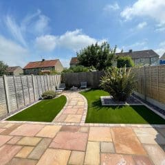 A backyard with a stone patio and curved pathway leads to artificial grass sections, enhanced by nearby block paving. Two lounge chairs are positioned near a tall plant. The area is surrounded by wooden fences, with houses and a cloudy sky in the background.