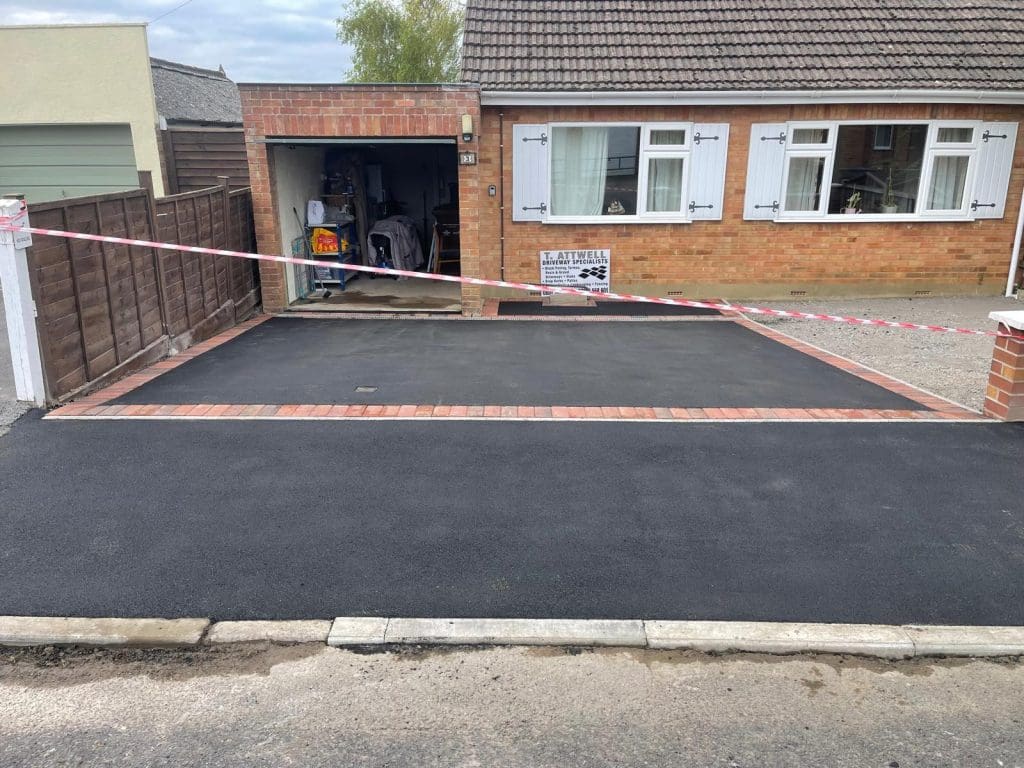 A freshly laid asphalt driveway installation graces the front of a one-story brick house with a garage. A red and white caution tape blocks the entrance. The driveway features a neat brick border, and the garage contains various items.