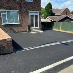 A freshly paved resin driveway in front of a brick house with large windows and a glass door. There's a small set of steps leading to the entrance. A green fence separates the yard from a neighboring house, and a streetlamp is visible on the left.