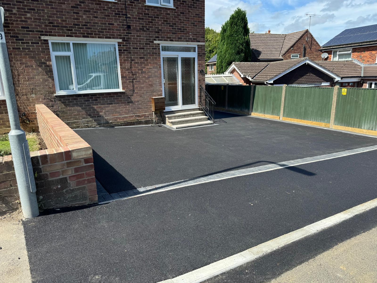 A freshly paved resin driveway in front of a brick house with large windows and a glass door. There's a small set of steps leading to the entrance. A green fence separates the yard from a neighboring house, and a streetlamp is visible on the left.