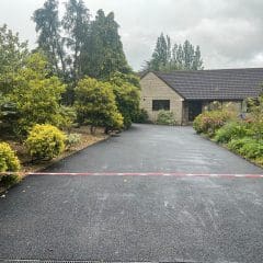 A house with a gray roof sits at the end of a long, freshly-paved resin driveway. Lush green shrubs and trees border the path, while a thin pink tape stretches across it. The sky is overcast, casting a moody light on this expertly crafted work by skilled driveway installers.