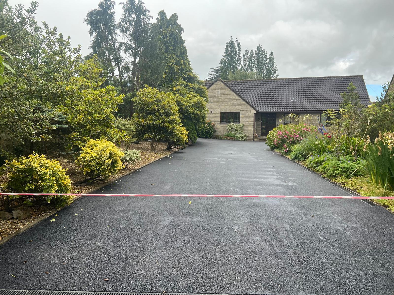 A house with a gray roof sits at the end of a long, freshly-paved resin driveway. Lush green shrubs and trees border the path, while a thin pink tape stretches across it. The sky is overcast, casting a moody light on this expertly crafted work by skilled driveway installers.