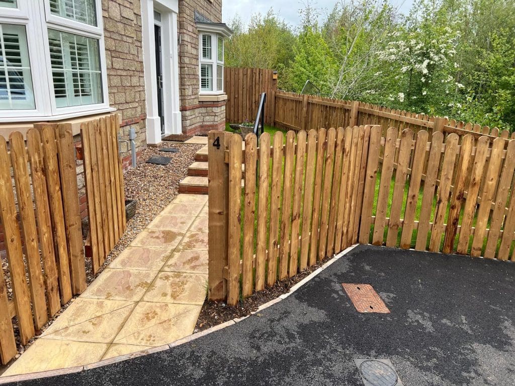 A wooden fence surrounds a small front yard with a paved walkway leading to the house's front door. The area, enhanced by resin bound stones, has some grass and small stones by the path. A lush, green background of trees is visible.