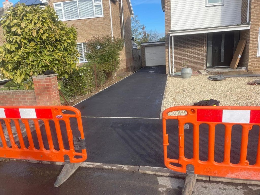 A newly paved black driveway leads to a garage beside a brick house, showcasing expert driveway installation. Two orange barriers block the entrance. A small gravel area is to the right, and a large bush with green leaves is on the left.