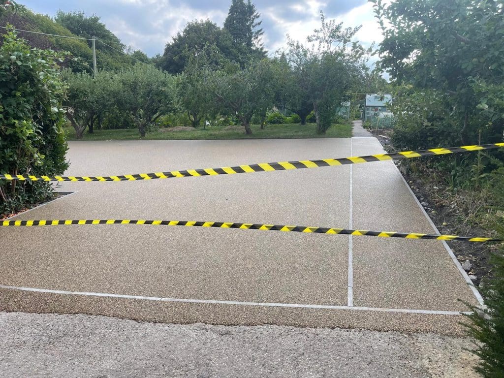 A freshly paved resin driveway is bordered with yellow and black caution tape. The path leads into a garden area with green trees and shrubs under a cloudy sky, showcasing the meticulous work of expert driveway installers.