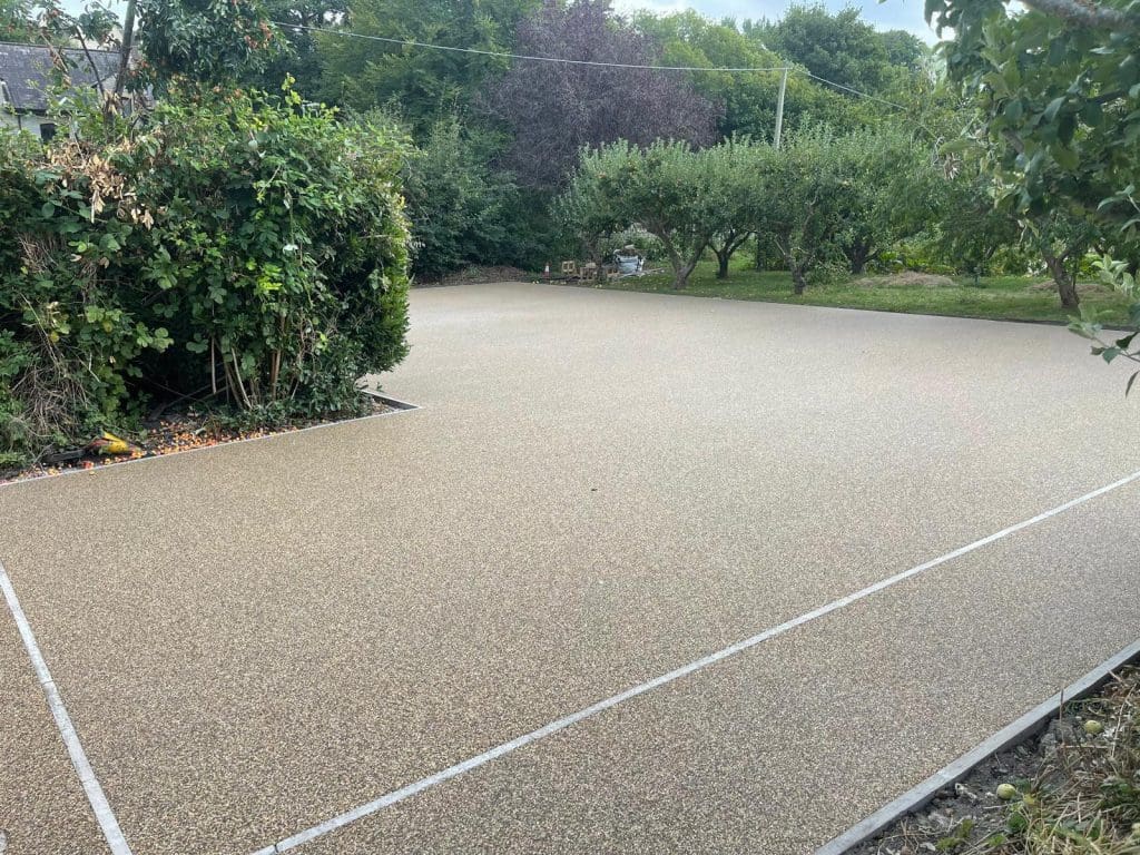 A new, smooth, beige resin patio with elegant white boundary lines is surrounded by lush greenery, including trees and bushes. This outdoor oasis in the garden area showcases the expertise of driveway installers specializing in block paving.