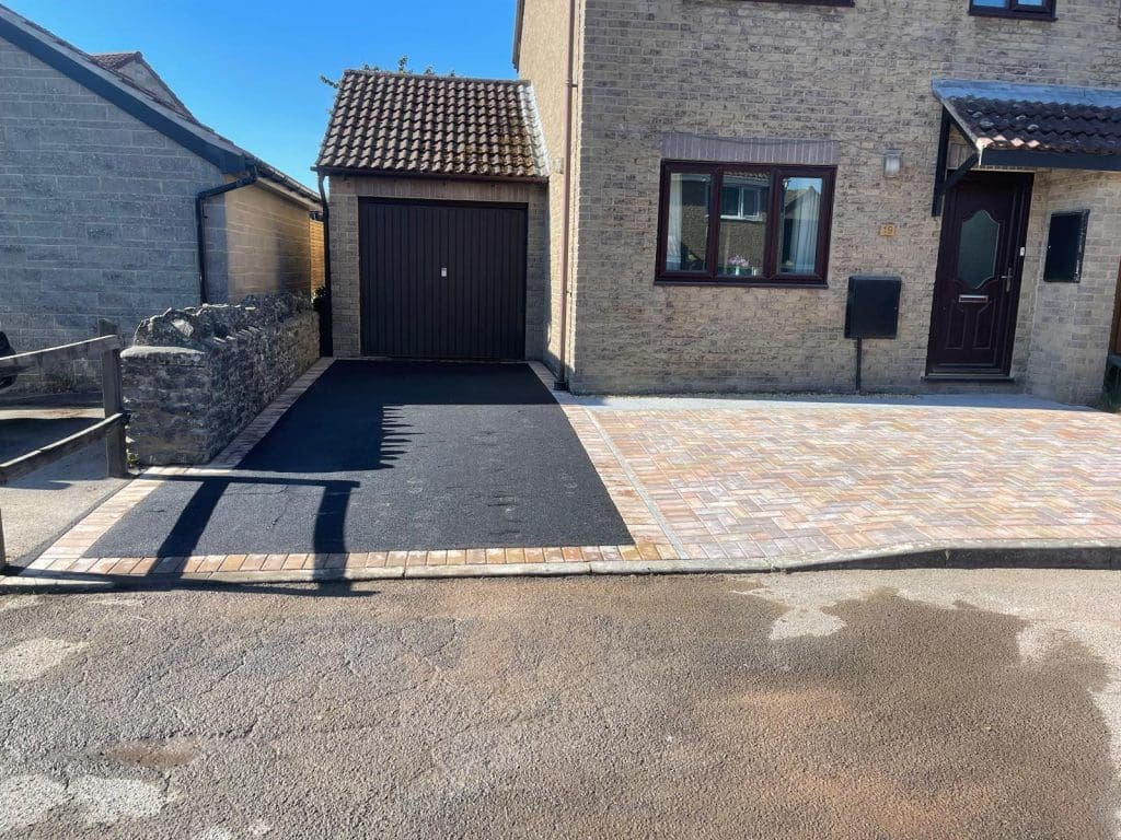 A paved driveway with a blend of dark asphalt and light-colored bricks leads to a garage attached to a stone house. Resin Installers created the elegant surface. There's a small wall on the left and a mailbox near the front door. Sunny weather casts shadows on the driveway.