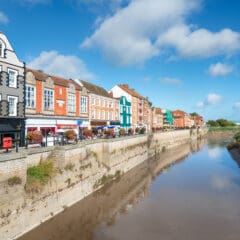 A calm river flows through a cityscape with colorful buildings lining the waterfront under a blue sky. The structures are mostly brick, with vibrant facades and multiple windows, creating a picturesque urban scene.