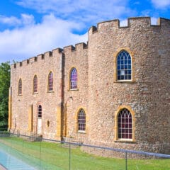 A stone castle with arched windows and a crenelated roof stands beside a glass walkway. The building is surrounded by lush greenery under a blue sky with scattered clouds.