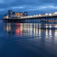 A pier with a building extends over the calm sea at dusk, illuminated by rows of bright lights. The water reflects the lights, creating a shimmering effect. The sky is a deep blue with scattered clouds, and the sandy beach is partially visible.
