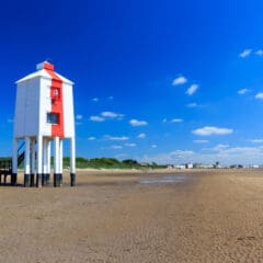 A white and red lighthouse stands on stilts on a sandy beach under a bright blue sky. The shoreline is visible in the background with green vegetation and distant buildings. Scattered clouds dot the sky.