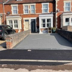 A freshly resurfaced driveway with light gray gravel in front of a red-brick townhouse, bordered by low brick walls. A black SUV is parked on a gravel area to the left, and the sidewalk in front is newly paved.