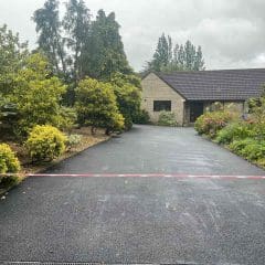 A smooth, newly paved asphalt driveway leads to a stone house with a dark roof, surrounded by lush green trees and bushes. A red and white barrier tape is stretched across the driveway entrance.