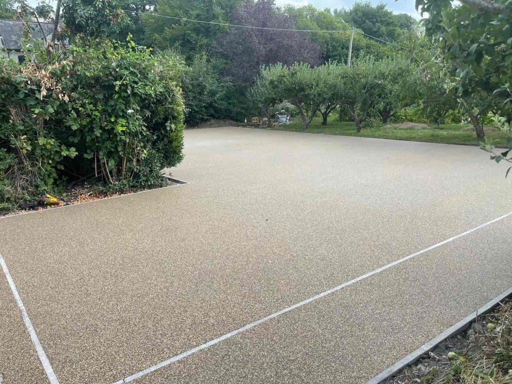 A freshly laid resin driveway or patio with a smooth, light-colored surface, bordered by greenery, bushes, and trees in a garden setting. Thin white lines outline sections of the paved area.