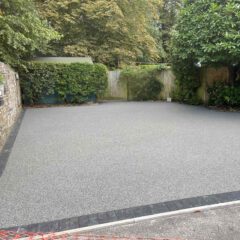 A freshly laid, smooth grey resin driveway bordered with dark bricks, surrounded by stone walls, green plants, and trees in a residential garden.